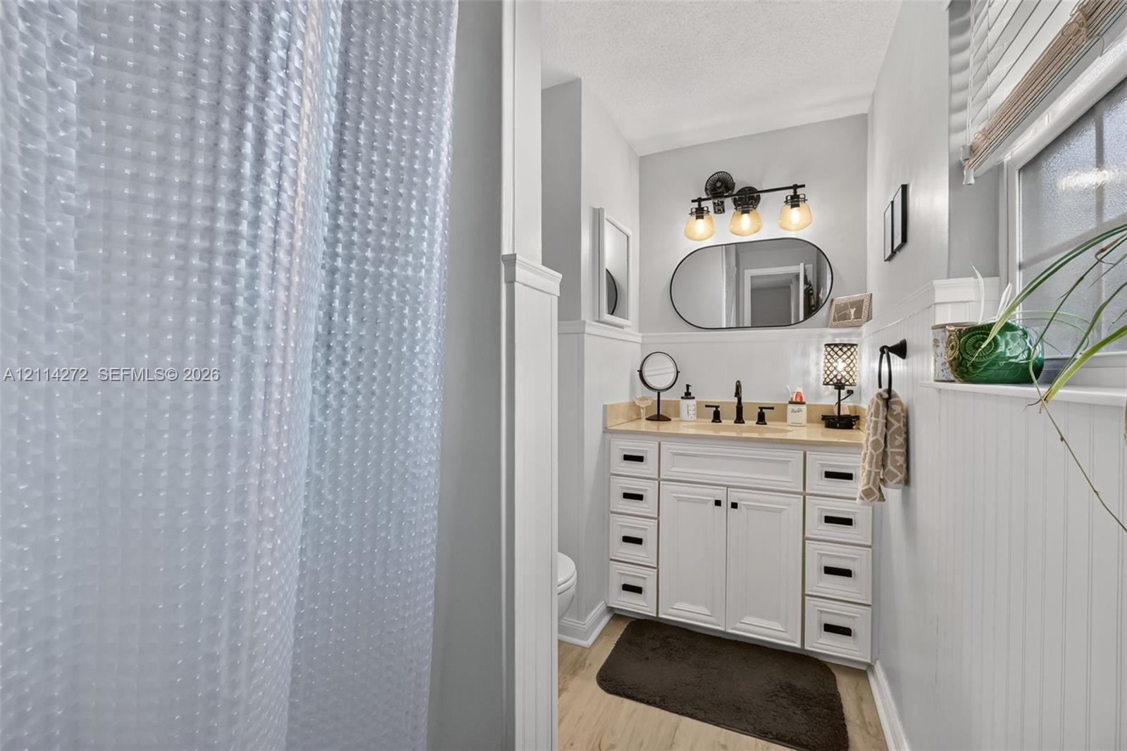 White vanity cabinet with multi-drawer storage, stone countertop, oval wall mirror, and three-light matte black fixture, complemented by wainscoting and wood-finish flooring