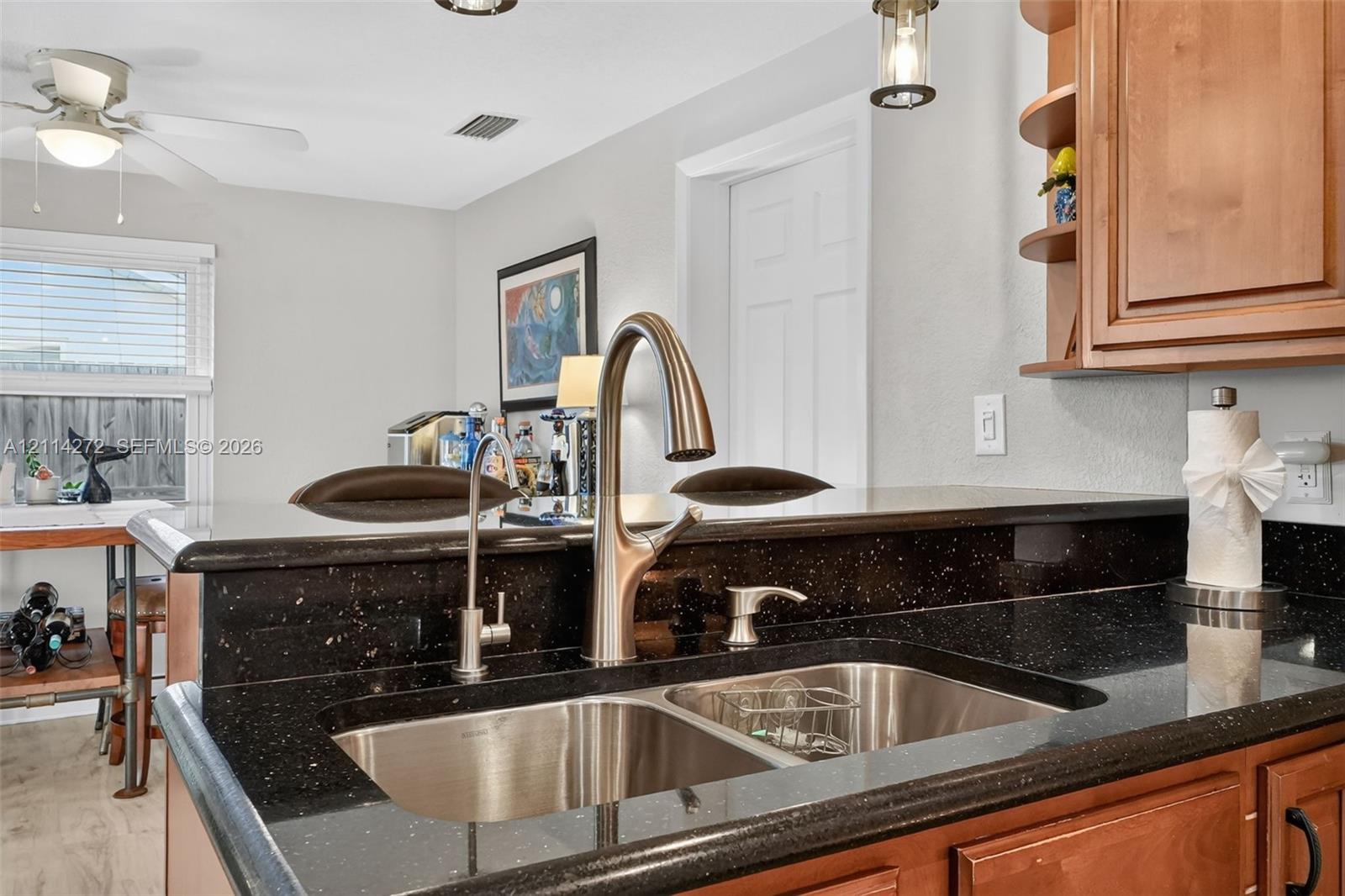 Kitchen area featuring dark stone countertops, an integrated dual-basin stainless steel sink, a brushed nickel high-arc faucet, wood cabinetry with open shelving, and pendant lighting