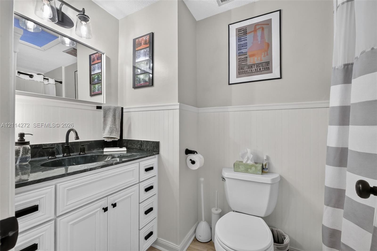 Bathroom featuring a vanity with dark stone countertop, integrated sink, white cabinetry with matte black hardware, and white beadboard wainscoting