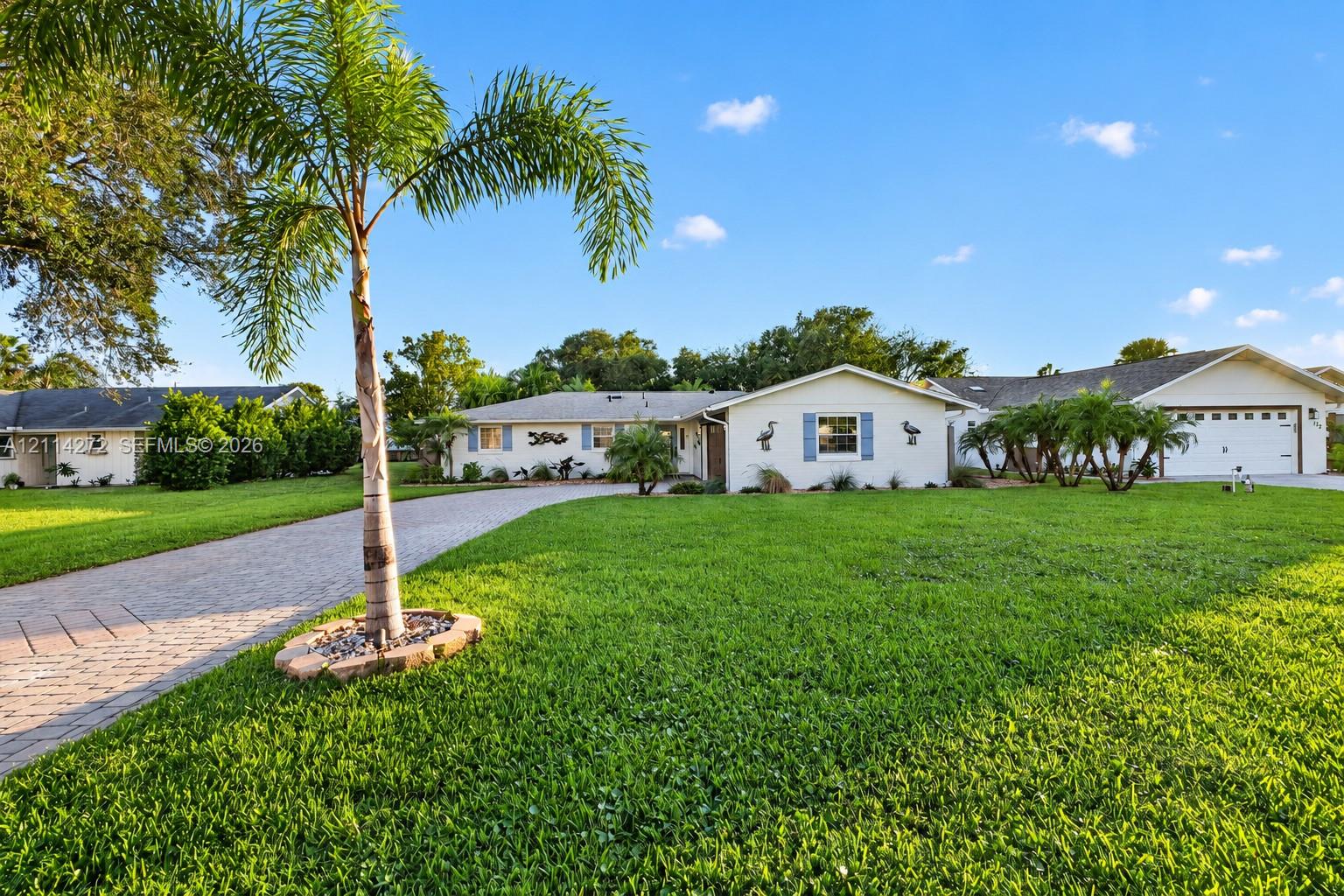 Single-story exterior featuring a paver driveway, manicured lawn, attached two-car garage, and mature palm landscaping