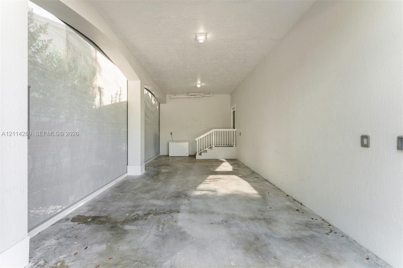 Covered parking area featuring textured concrete flooring, white stucco walls, overhead lighting, and built-in white railings