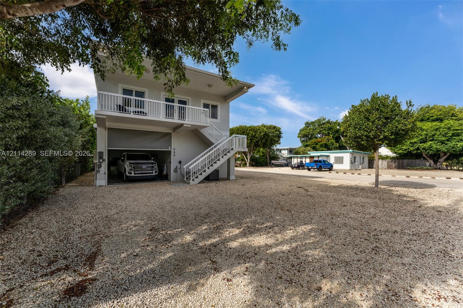 Two-story exterior featuring a gravel driveway, an open-concept ground-level garage, and a second-story balcony with white railing and external staircase access