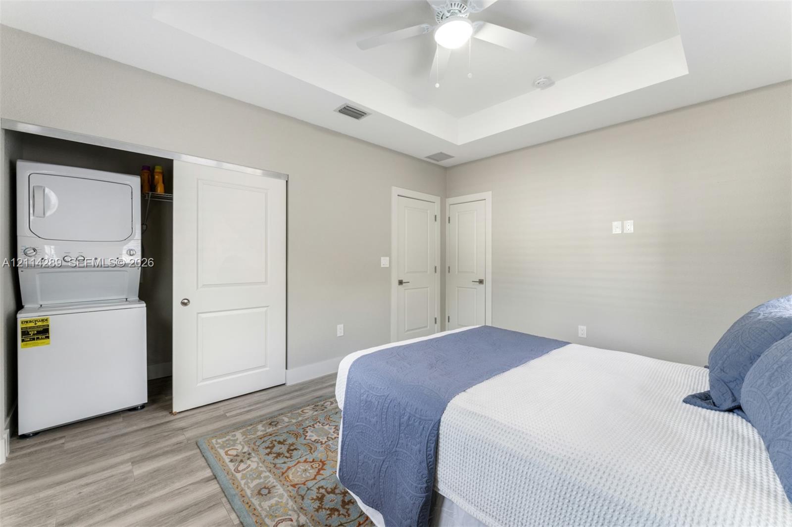 Tray ceiling featuring a central light-integrated ceiling fan, wood-finish flooring, and built-in stacked laundry unit