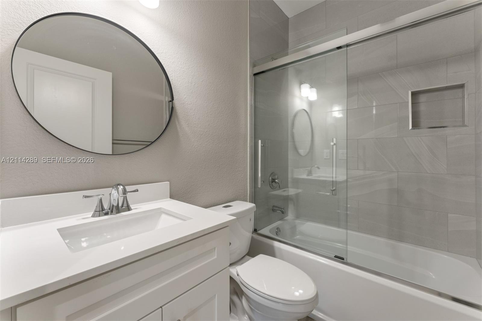 Bathroom featuring a vanity with a white countertop and rectangular undermount sink, oval wall mirror, sliding glass shower door, and tile-surround tub with a built-in recessed niche