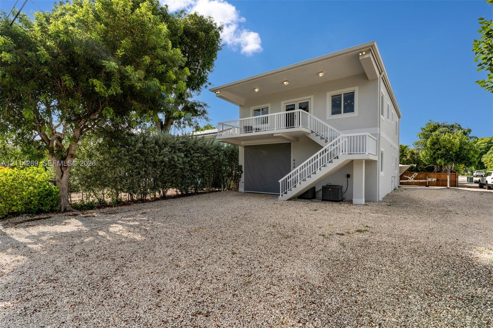 Two-story exterior featuring a gravel driveway, elevated balcony with white railing, external staircase, recessed soffit lighting, and an expansive garage door
