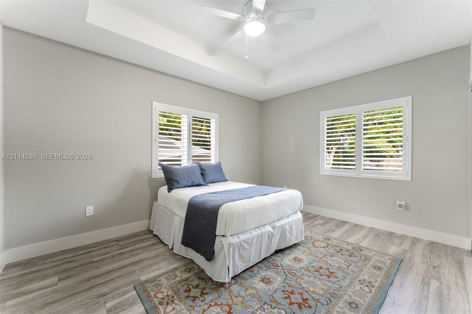 Tray ceiling with a central white ceiling fan, wood-finish flooring, and white plantation shutters on dual windows