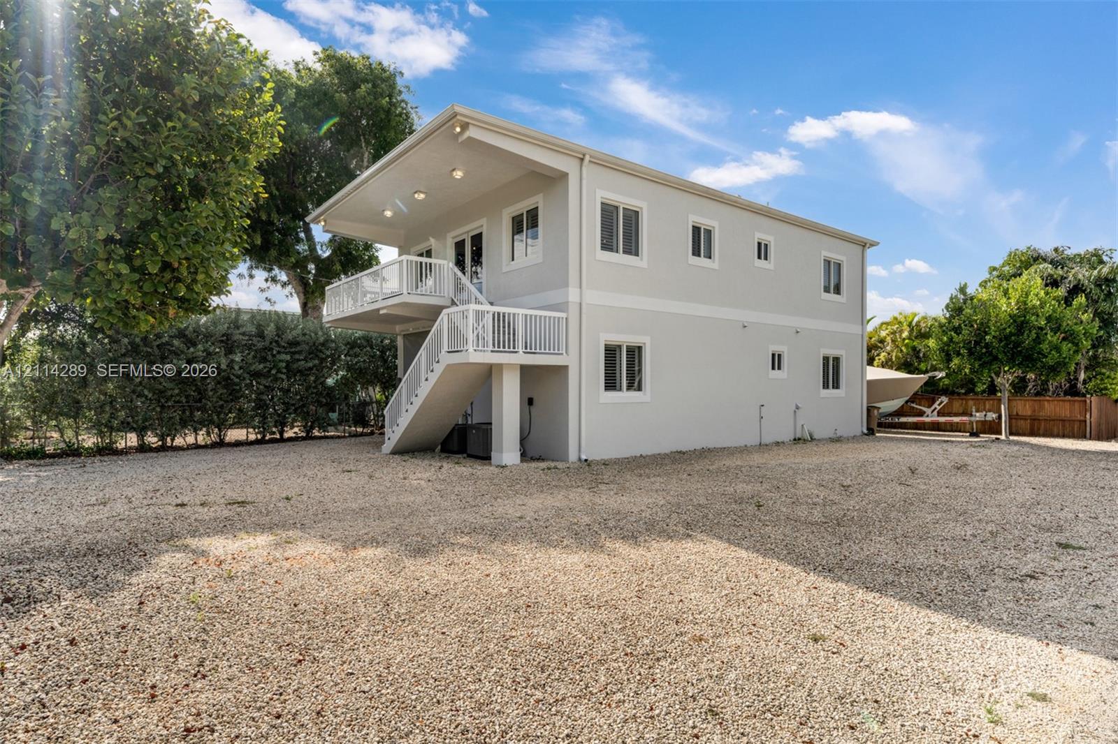 Two-story structure featuring light-toned smooth siding, a raised upper-level deck with white railings, an exterior staircase, and a sprawling gravel grounds area