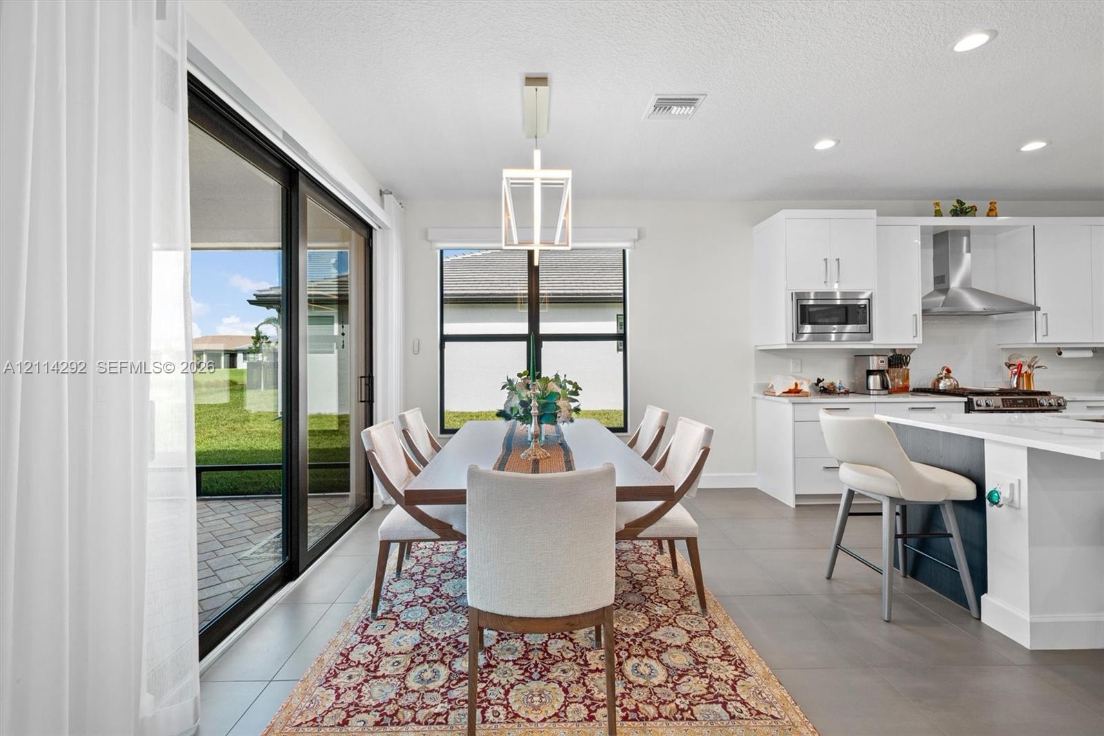 Open-concept kitchen featuring white shaker-style cabinetry, stainless steel appliances, a central island, and large-format tile flooring, complemented by sliding glass doors leading to a paved patio area