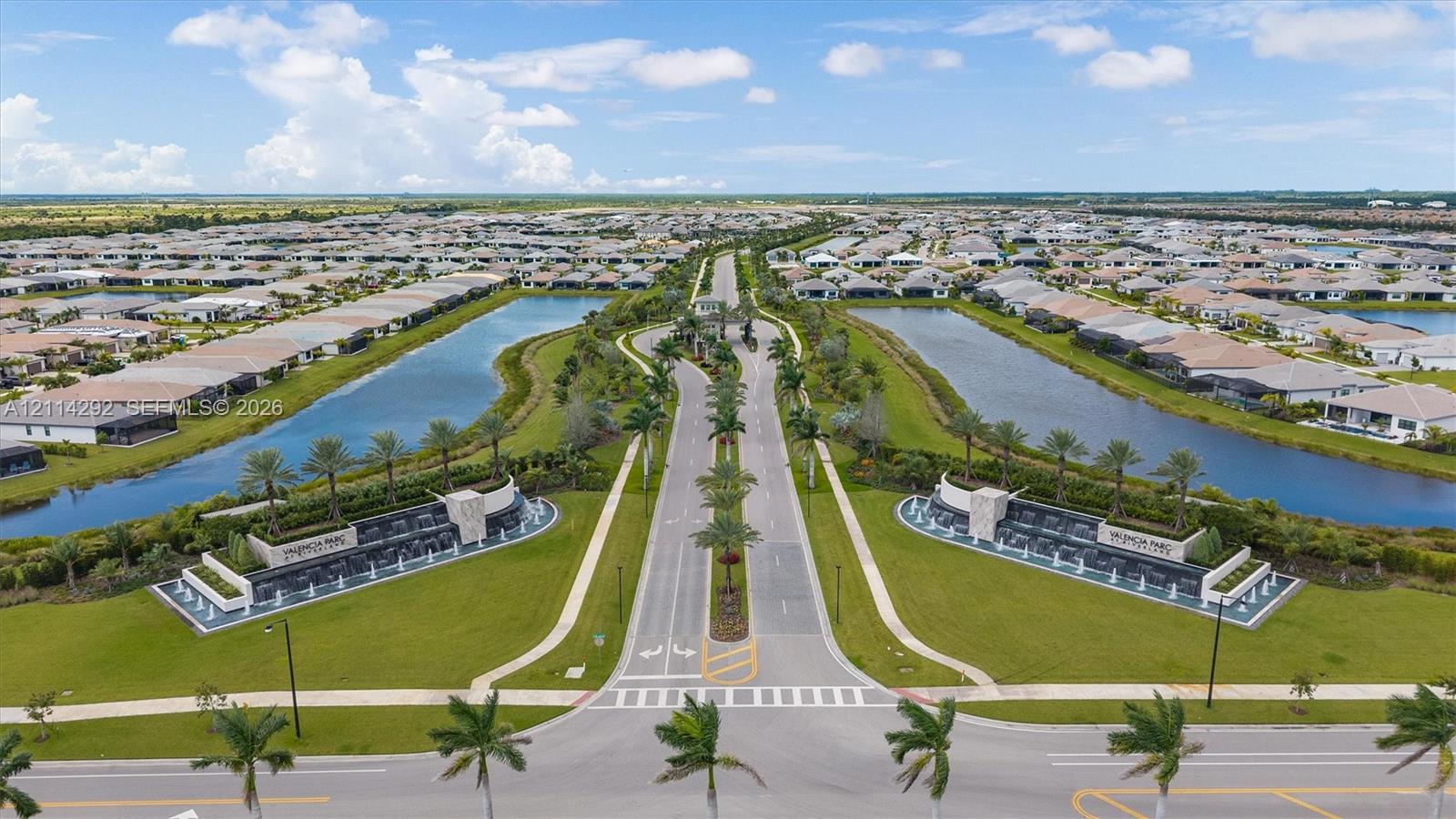 Aerial perspective of community entrance featuring tiered water fountains, stone-clad monument signage, manicured landscaping, palm tree-lined boulevards, and expansive retention ponds