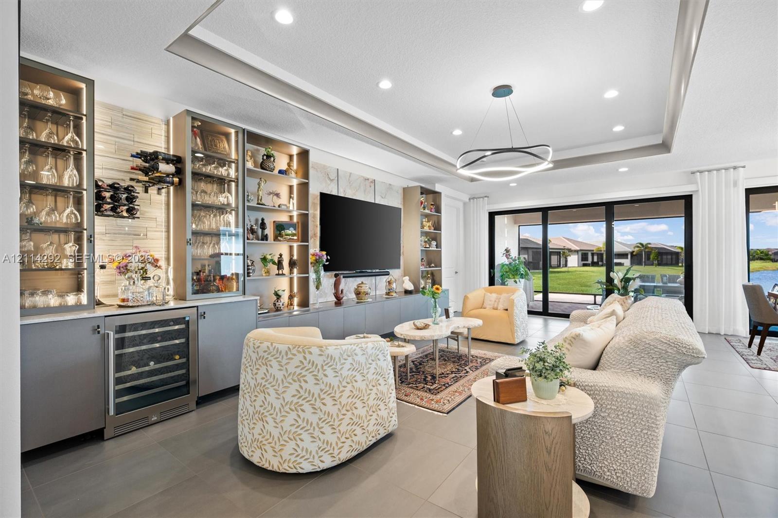 Custom built-in cabinetry with integrated glass-front display shelving and beverage refrigerator, tray ceiling with recessed lighting, large-format tile flooring, and multi-panel sliding glass doors