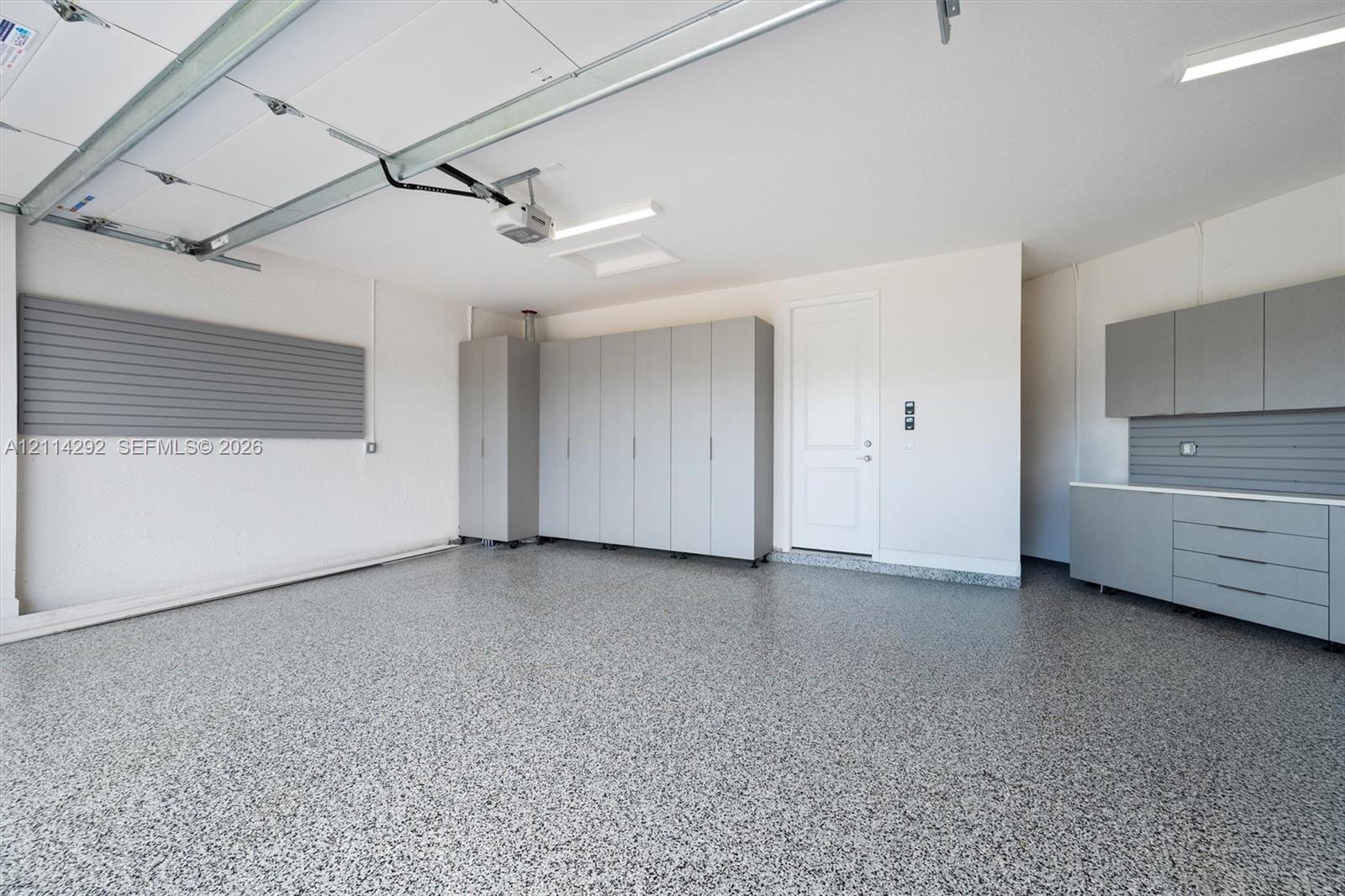 Finished garage featuring speckled epoxy flooring, built-in grey cabinetry, wall-mounted storage panels, and overhead LED lighting