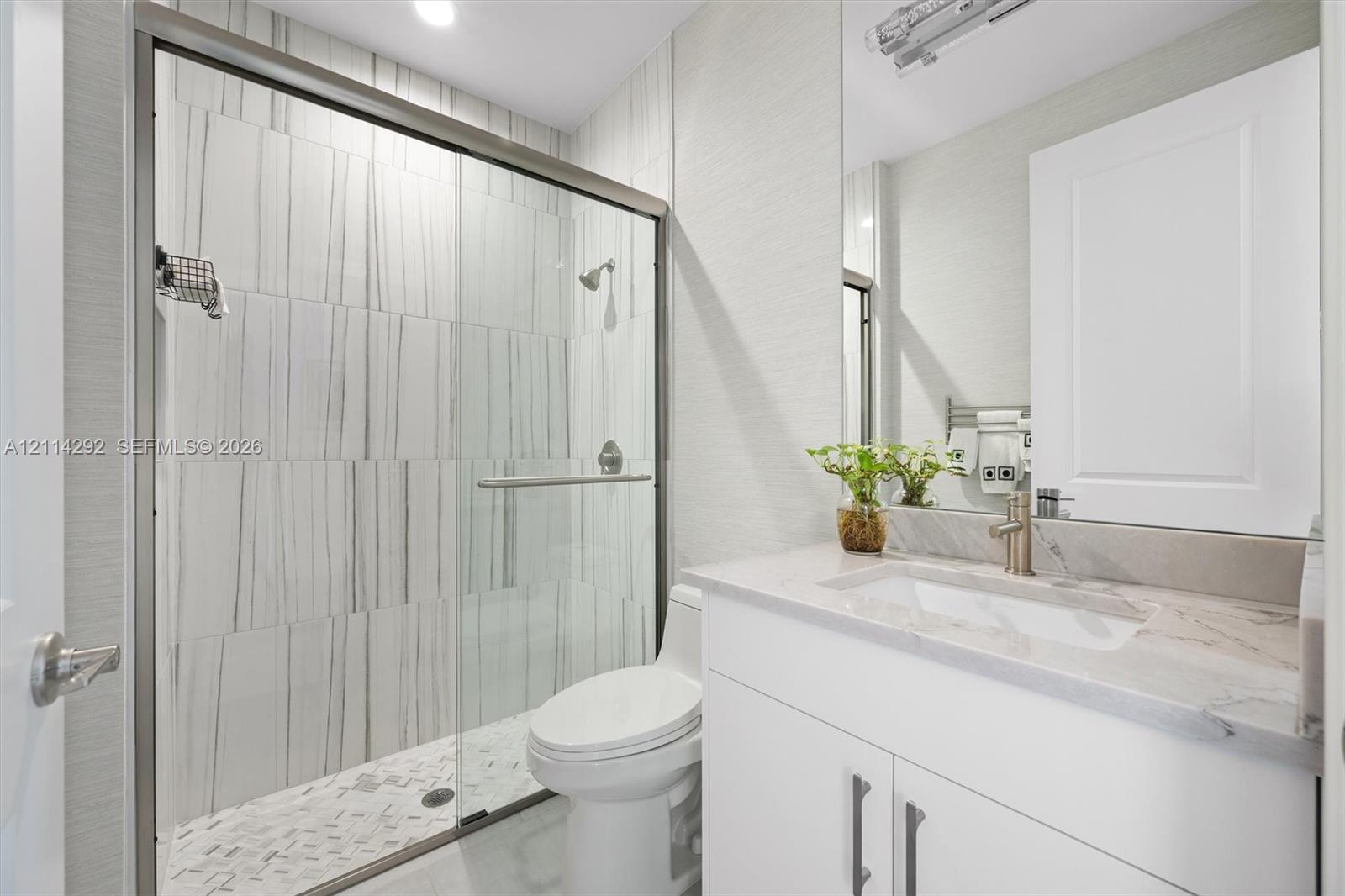 Bathroom featuring a walk-in glass shower with vertical-patterned wall tile, stone-surface countertop, integrated rectangular sink, vanity with white cabinetry, and brushed-metal hardware