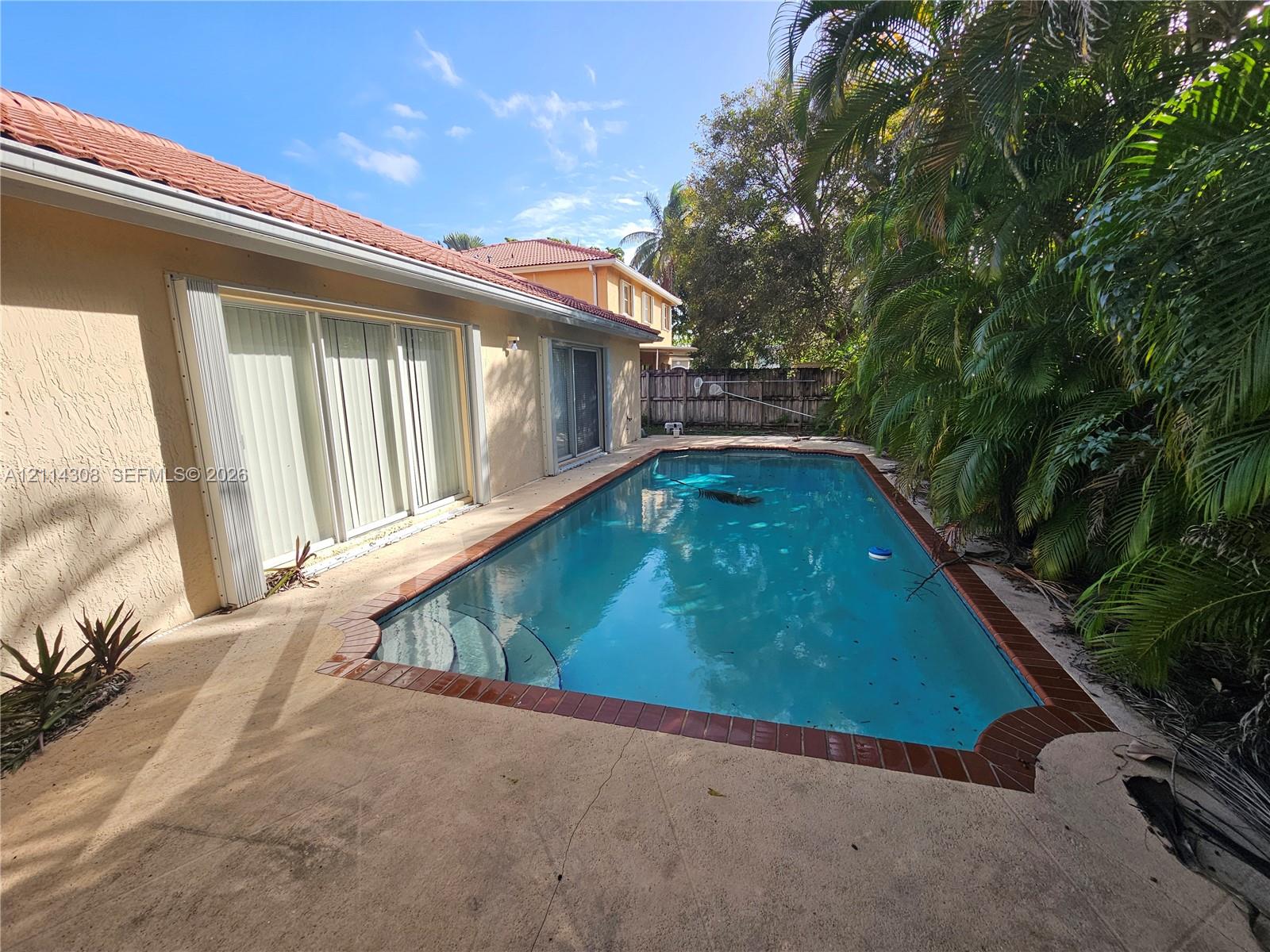 In-ground swimming pool with brick coping, concrete patio, sliding glass door access, and mature palm landscaping