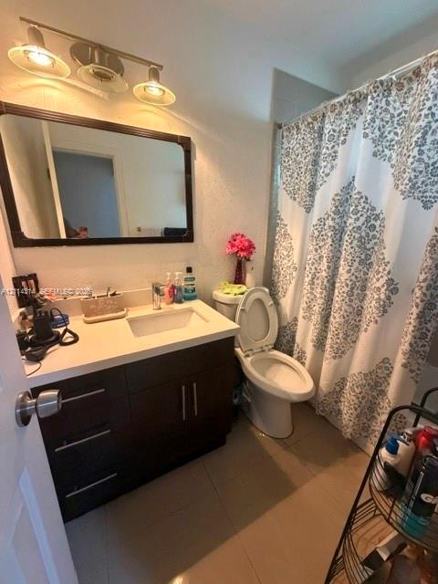 Bathroom featuring a vanity with a white countertop and rectangular undermount sink, dark-toned cabinetry with modern hardware, a rectangular wall-mounted mirror, three-light vanity fixture, and tiled flooring Bathroom featuring a vanity with a white countertop and rectangular undermount sink, dark-toned cabinetry with modern hardware, a rectangular wall-mounted mirror, three-light vanity fixture, and tiled flooring