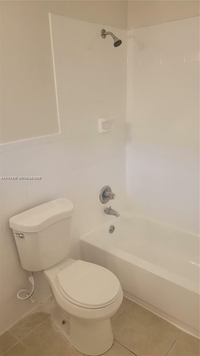 Full bathroom featuring a white porcelain toilet, a soaking tub with integrated shower head, tiled wall surround, and neutral-toned floor tile