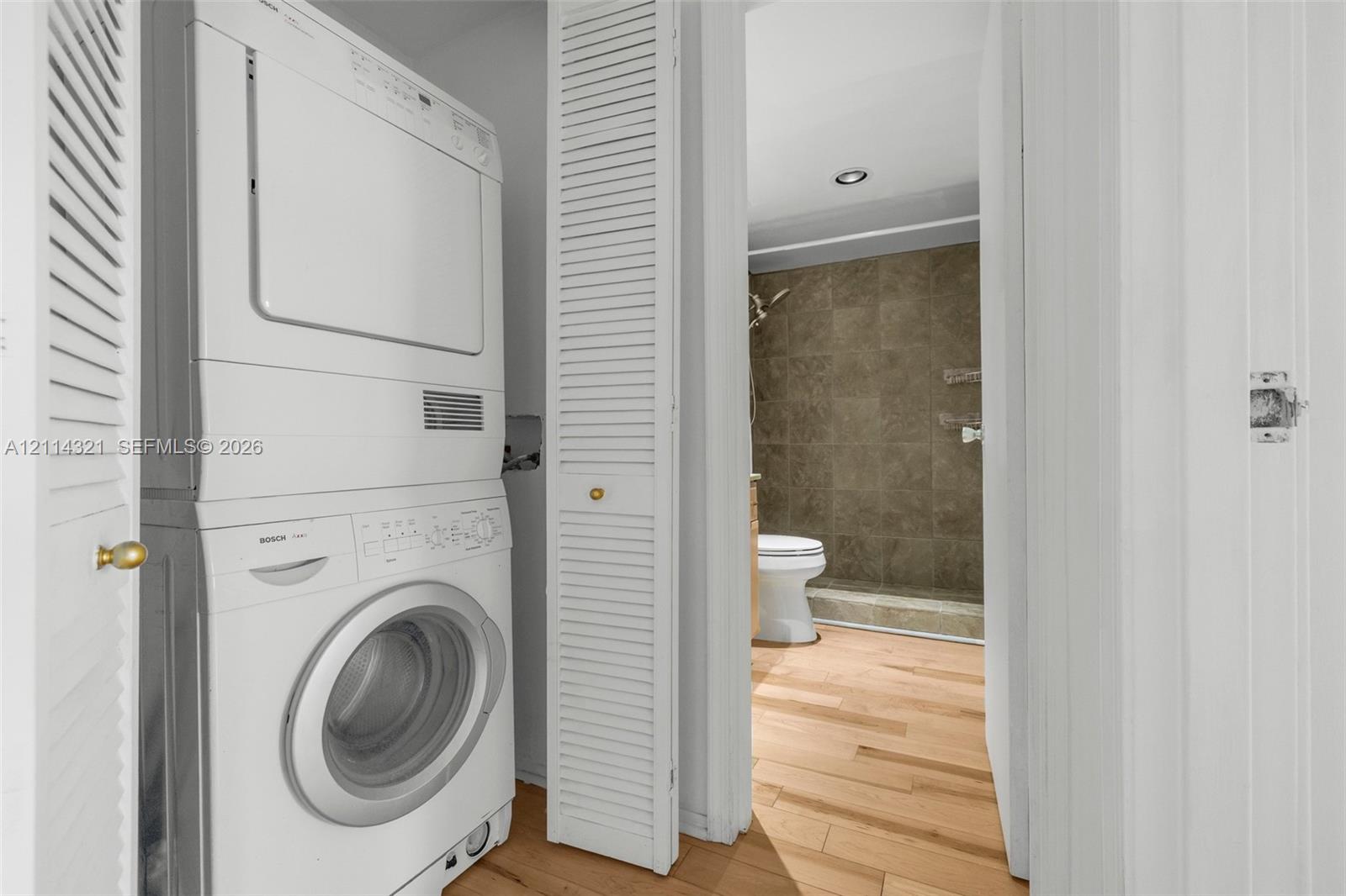 Stacked washer and dryer unit concealed behind white louvered doors, featuring wood-finish flooring and recessed ceiling lighting