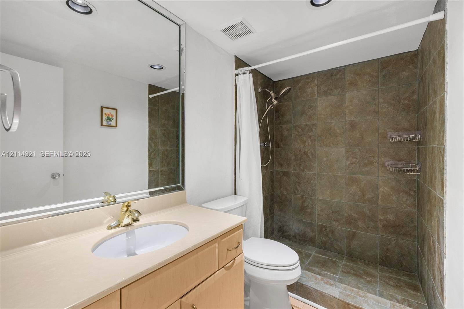 Bathroom featuring a vanity with a light-tone finish, solid surface countertop with an integrated oval basin, large wall-mounted mirror, recessed lighting, and a shower enclosure tiled with earth-toned stone
