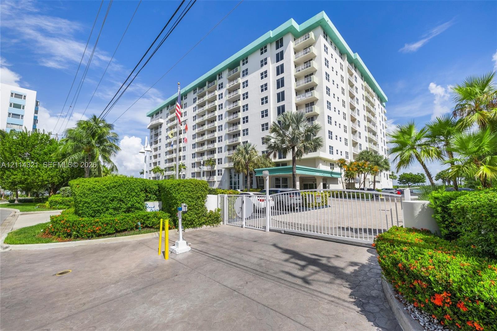 High-rise residential exterior featuring a gated entry, concrete-paved driveway, multi-level balconies, and mature tropical landscaping