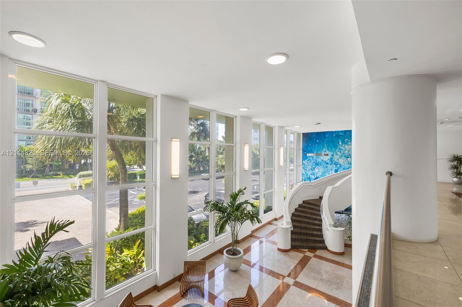 Floor-to-ceiling windows, sculptural curved staircase, stone flooring with decorative inlay, wall-mounted lighting fixtures, and high ceilings