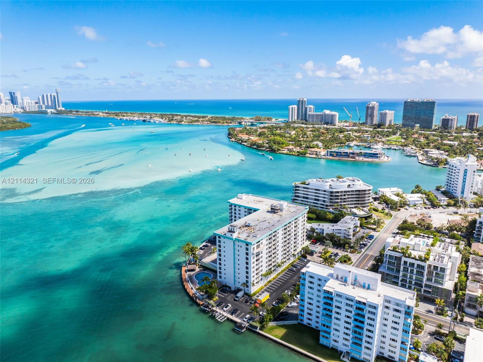 Aerial perspective of a multi-story residential building situated on a waterfront peninsula featuring a swimming pool, paved surface parking, and coastal water surroundings