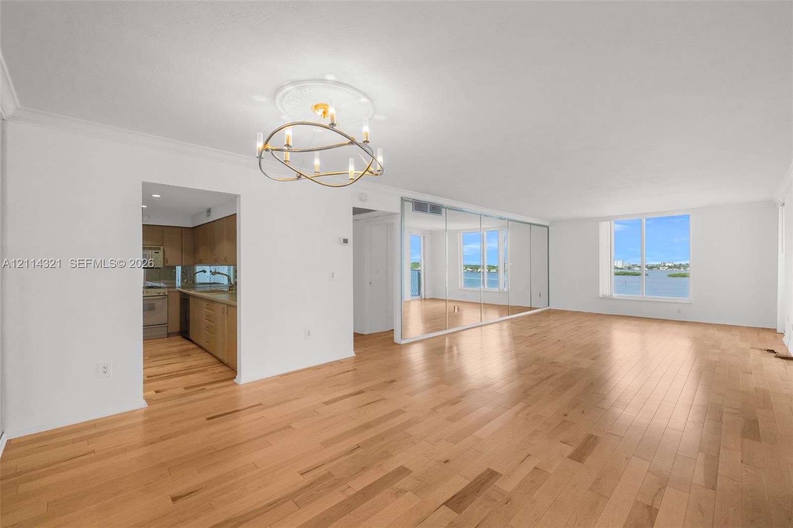 Open-concept living space featuring wood-finish flooring, a modern multi-bulb chandelier, mirrored wall paneling, and large windows with water scenery