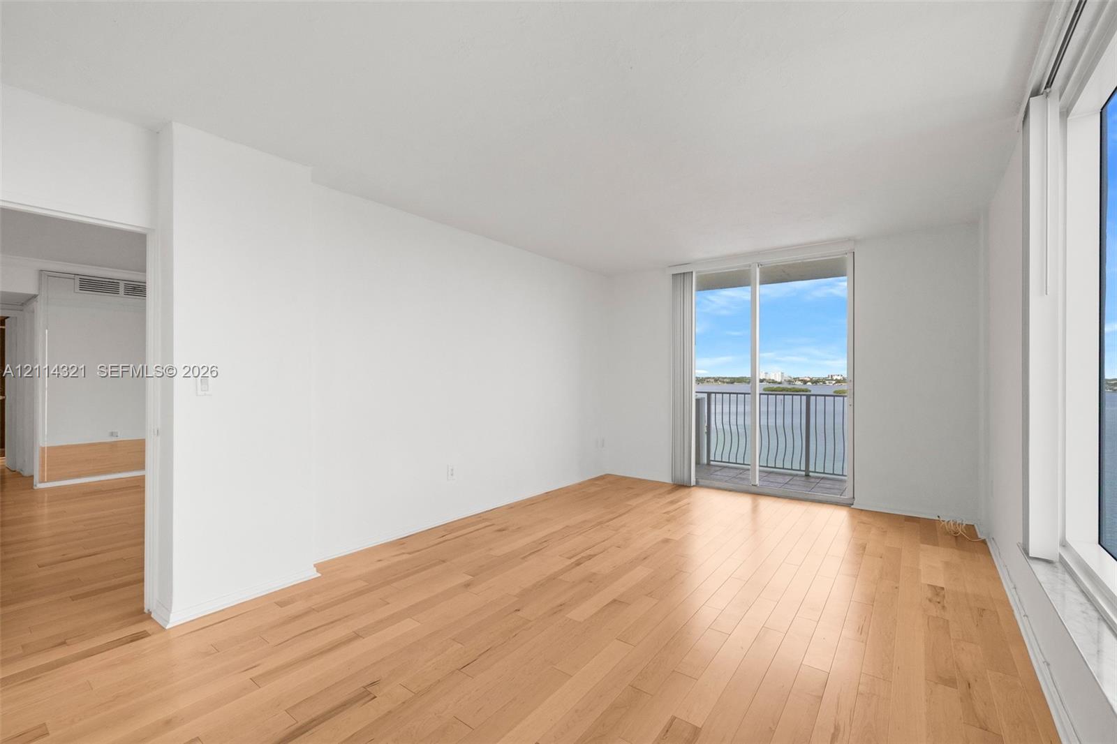 Living area featuring light-toned wood-finish flooring, floor-to-ceiling windows, and a glass sliding door opening to a private balcony with water scenery