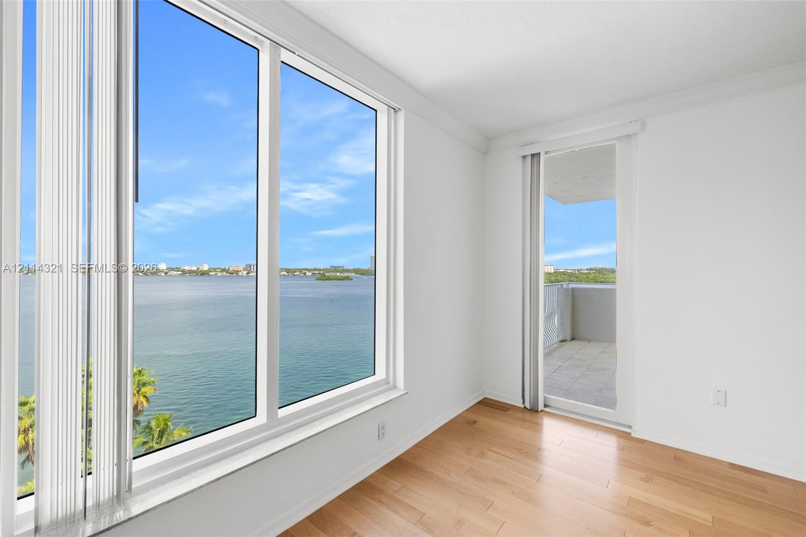 Large, wall-to-ceiling windows, wood-finish flooring, and a sliding glass door leading to a private balcony overlooking the water