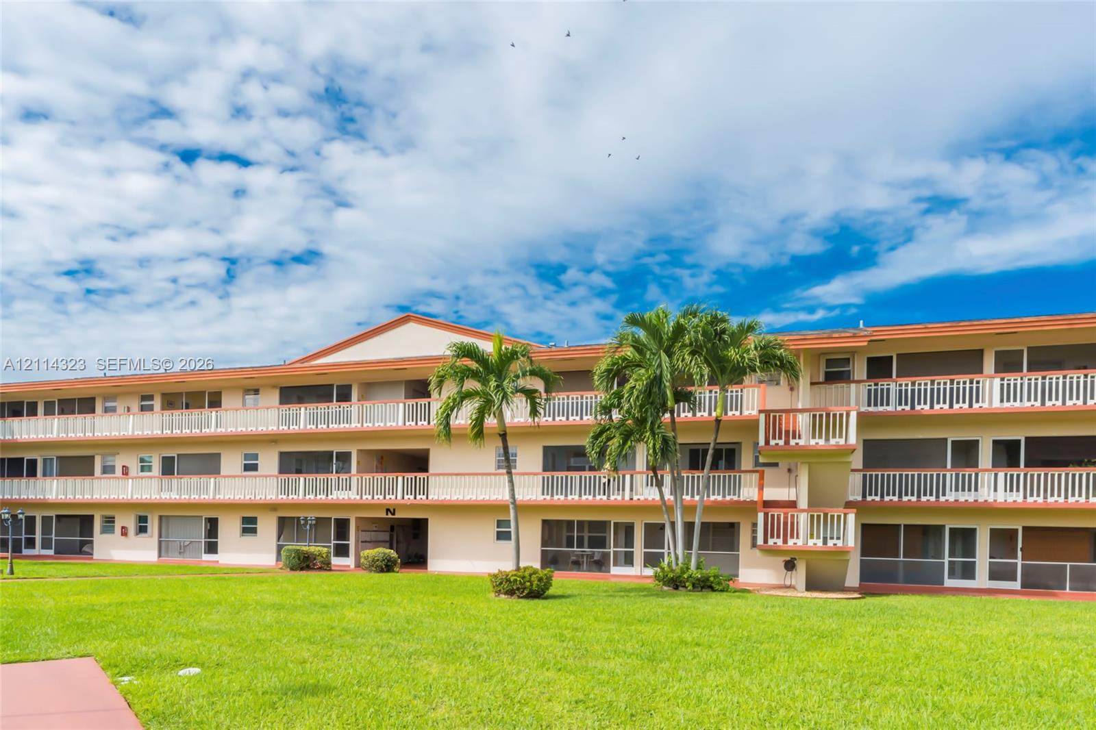 Three-story multi-family exterior featuring stucco siding, wrap-around balconies with white railings, screened-in exterior entryways, and mature palm trees set against a manicured lawn
