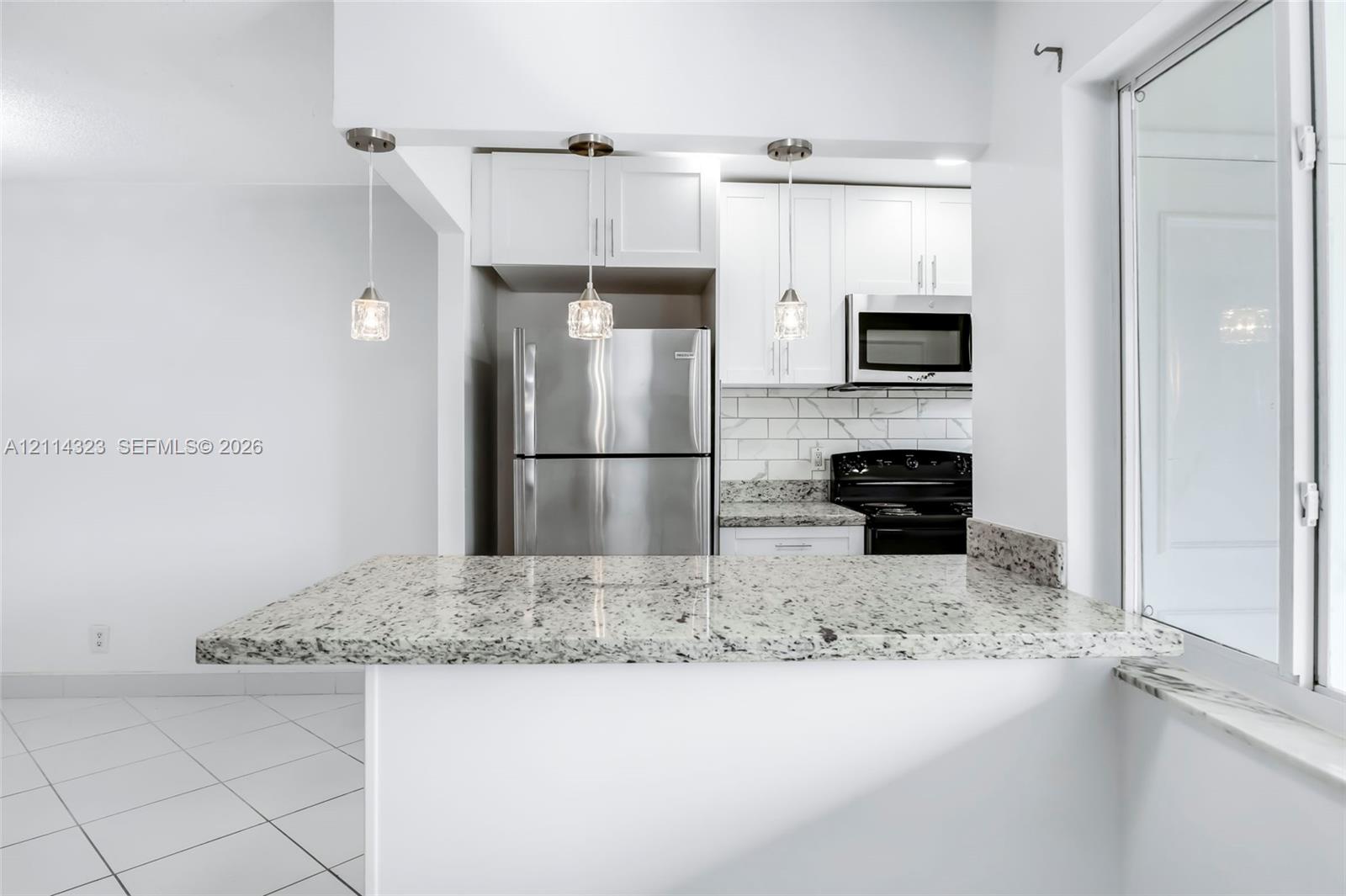 Kitchen featuring a peninsula with stone-finish countertop, three glass pendant lights, white shaker-style cabinetry, stainless steel appliances, and subway tile backsplash