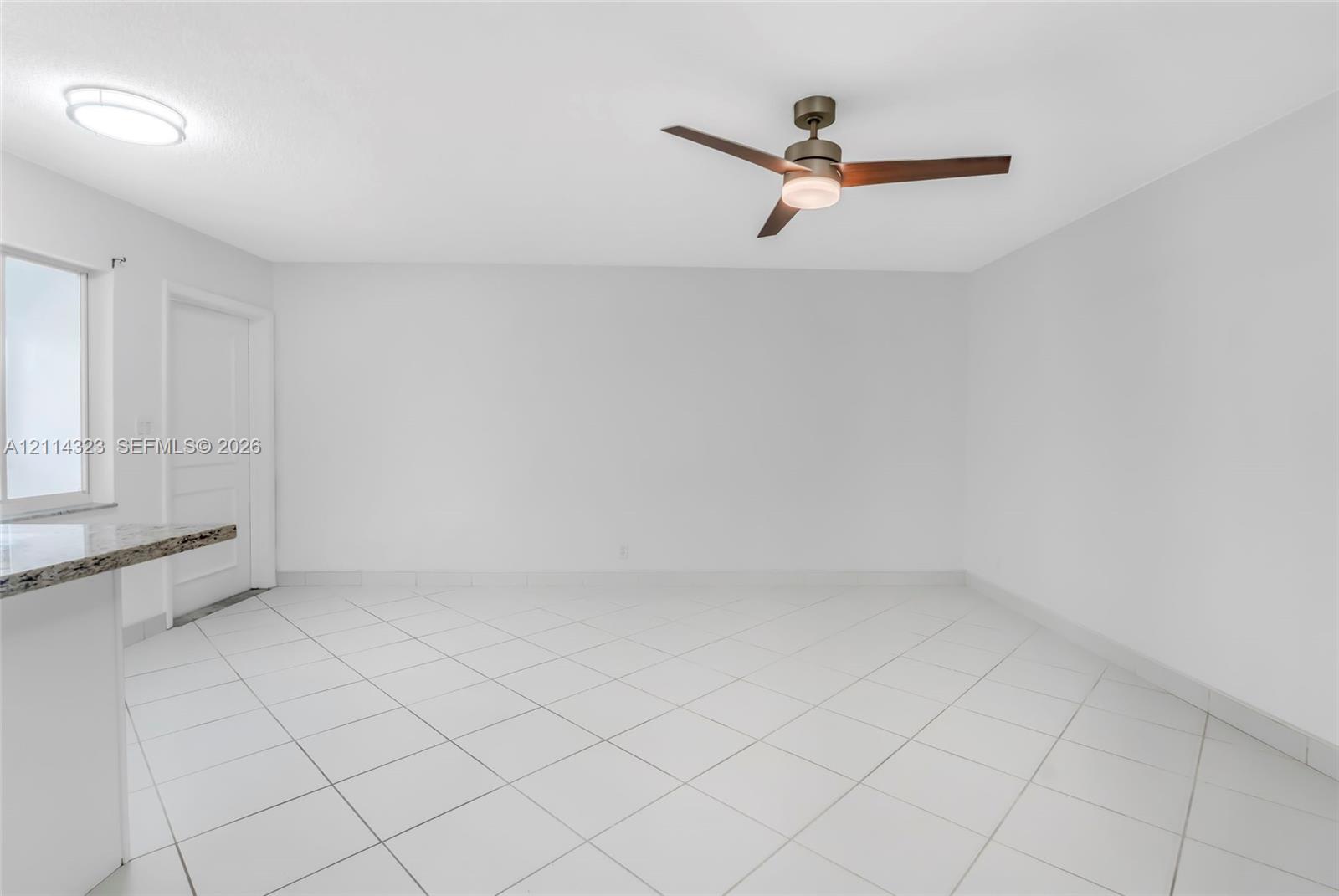 Neutral interior featuring large-format diagonal tile flooring, a modern three-blade ceiling fan with integrated lighting, a flush-mount circular ceiling fixture, and a granite-topped peninsula