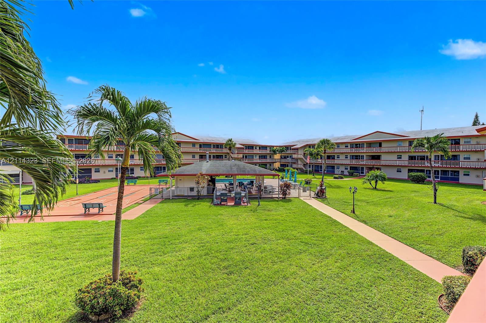 Community courtyard featuring a manicured lawn, a central pool pavilion, a shuffleboard court, and mature palm trees