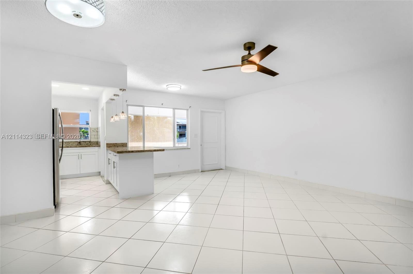 Open floor plan featuring white tile flooring, a kitchen peninsula with stone-finish countertops, three pendant lights, and a ceiling fan with integrated lighting