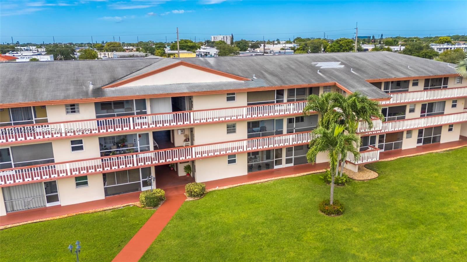 Multi-story residential building featuring covered private balconies with railings, exterior walkways, a gabled roofline, and expansive surrounding lawn areas with mature palms