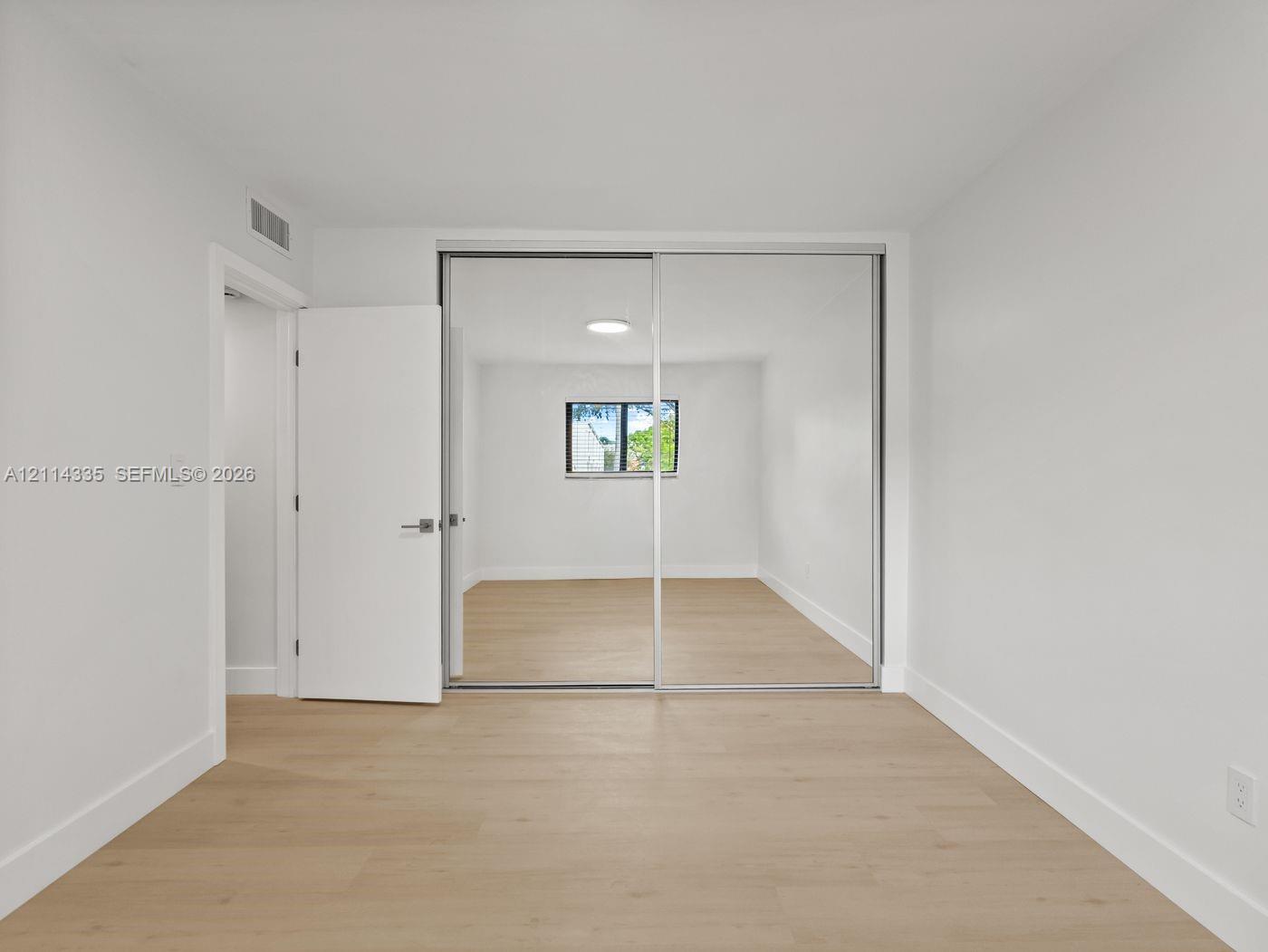 Light-toned wood-finish flooring, floor-to-ceiling sliding mirror closet doors, flush-mount ceiling lighting, and a single-pane window