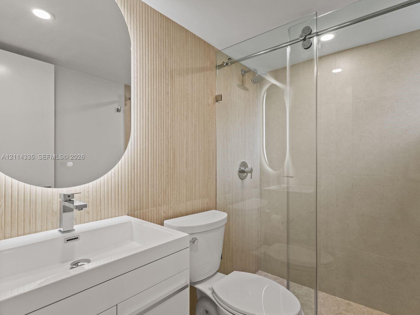 Bathroom featuring a white floating vanity, ribbed wood-panel accent wall, integrated circular mirror with backlighting, and a glass-enclosed shower with polished metal hardware