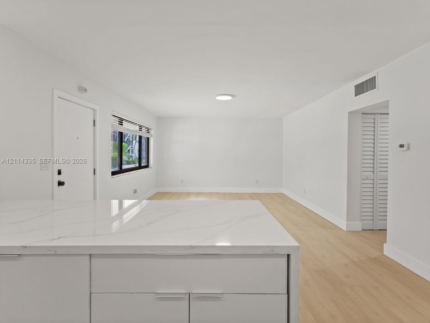 Open-concept interior featuring light wood-finish flooring, white cabinetry with stone-finish countertops, and a window with black trim