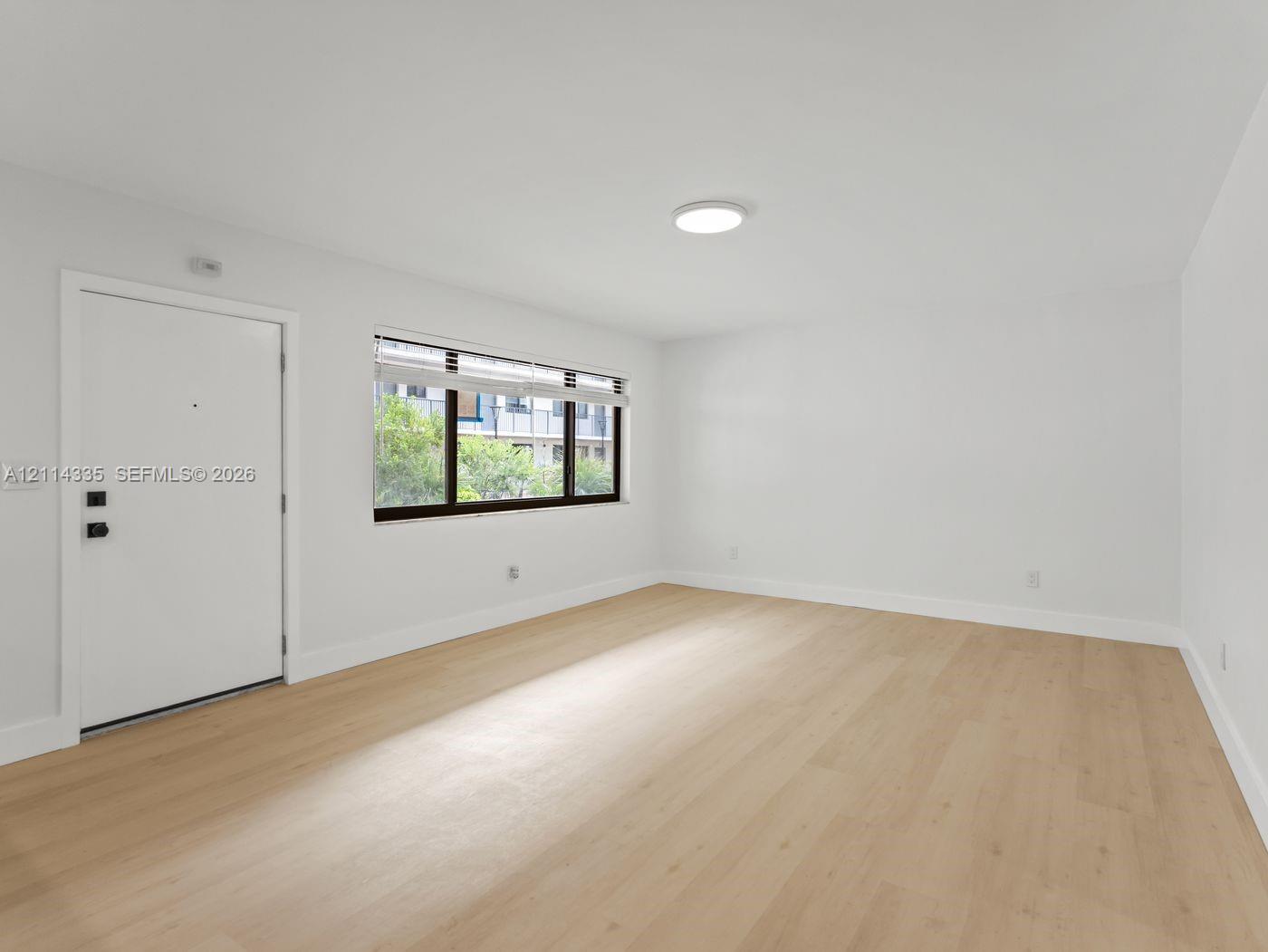 Neutral-toned space featuring wood-finish flooring, a flush-mount ceiling light, a white entry door with matte black hardware, and a window with a dark frame