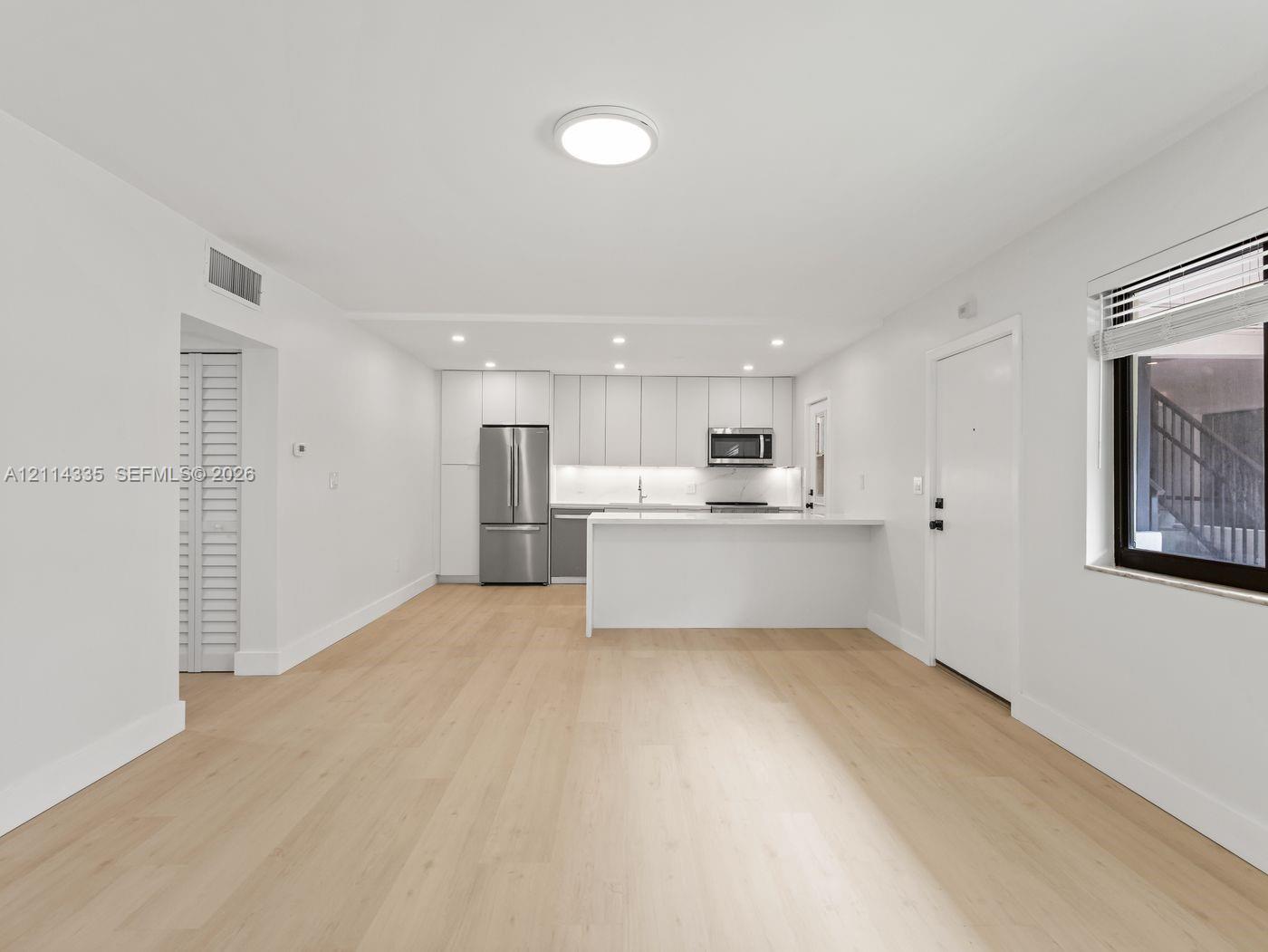 Open-concept area featuring light wood-finish flooring, white flat-panel cabinetry, a stainless steel refrigerator, a built-in microwave, and a white breakfast bar