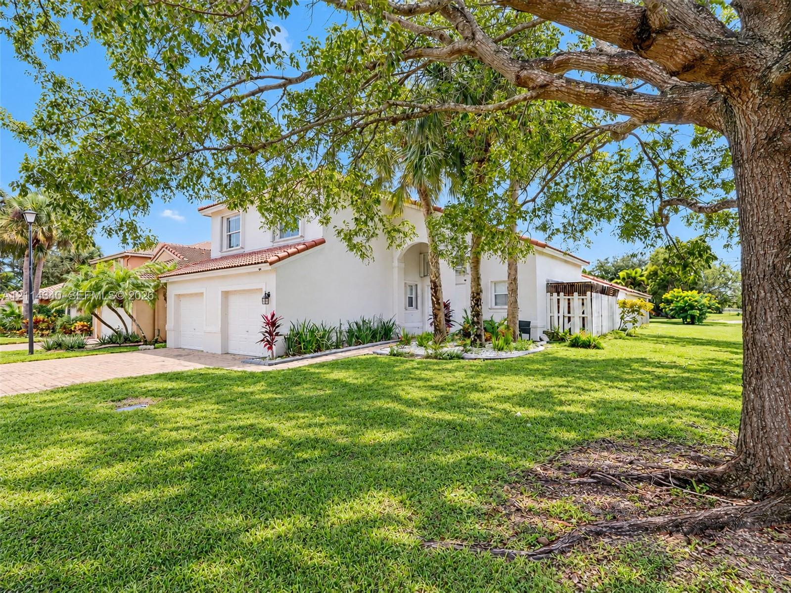 Stucco exterior featuring a red tile roof, two-car garage, paver driveway, and mature landscaping