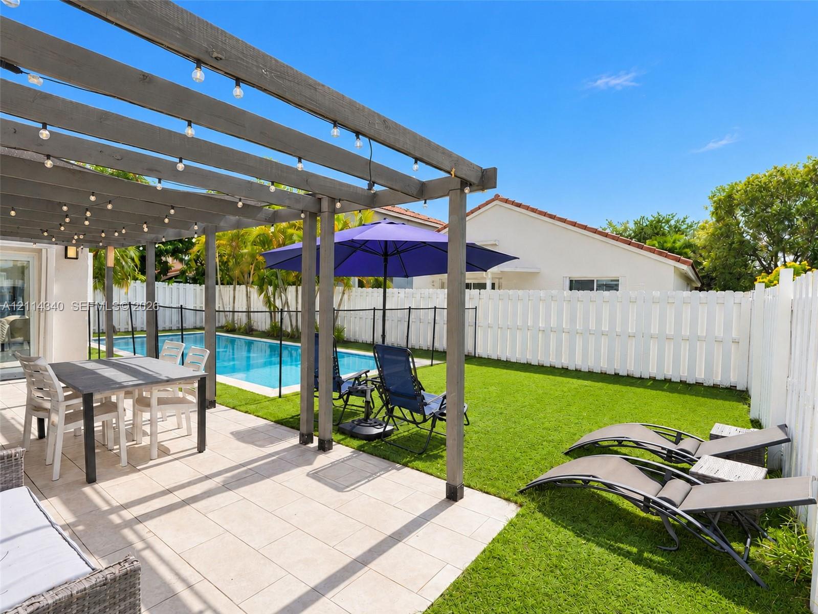 Outdoor living space featuring a wood-beam pergola, light-toned stone patio, private swimming pool, synthetic lawn, and perimeter privacy fencing
