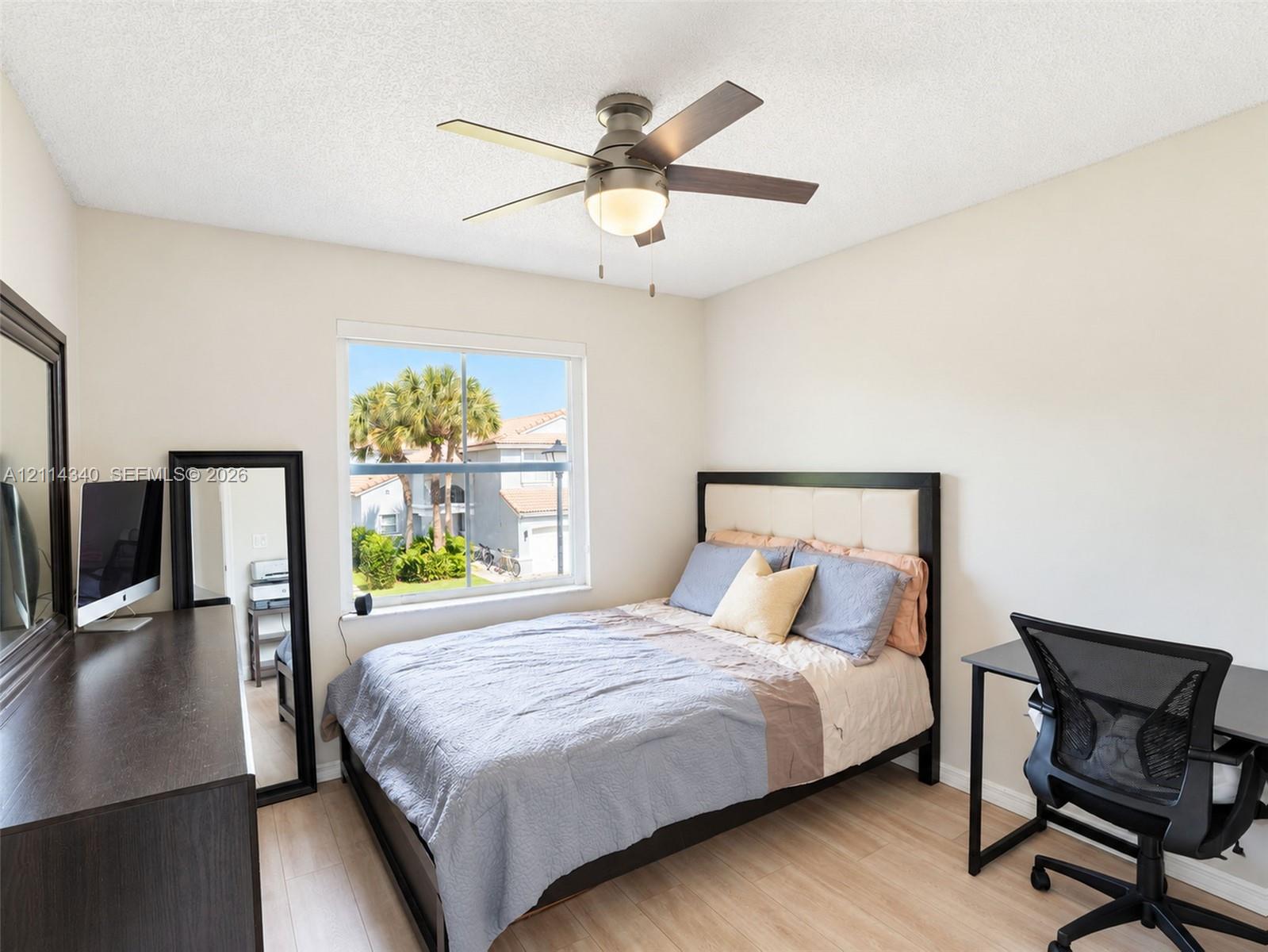 Light-filled room featuring wood-finish flooring, a central ceiling fan with integrated lighting, and a single-pane window with exterior palm tree scenery