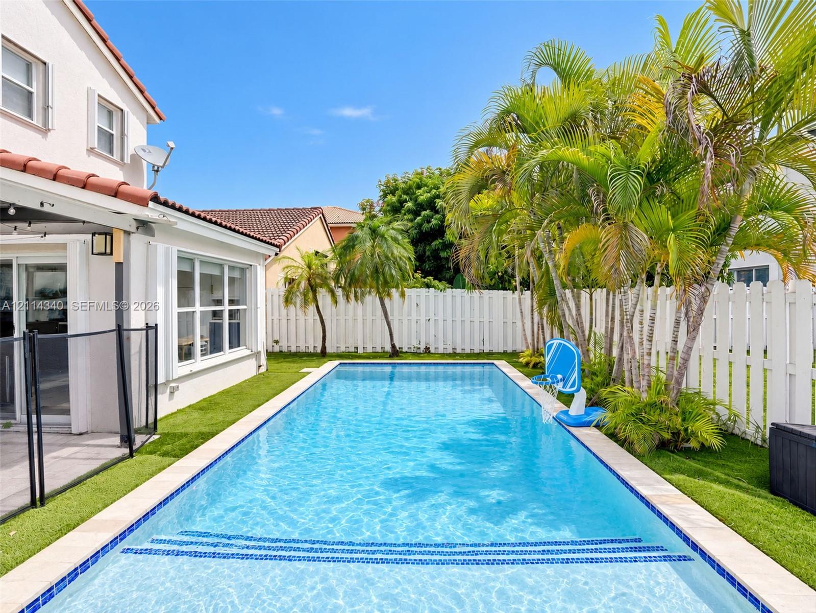 Rectangular in-ground pool with stone coping and built-in steps, surrounded by artificial turf and mature palm trees, enclosed by a white privacy fence
