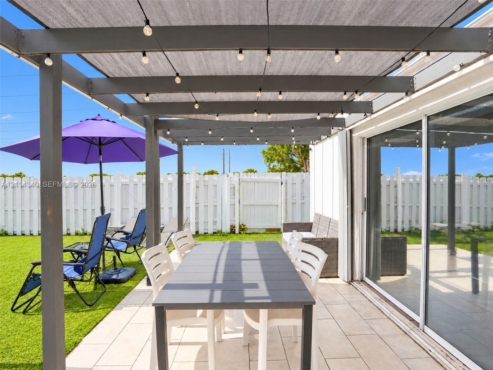 Covered patio featuring a grey wooden pergola, tile flooring, and integrated string lighting, complemented by a white privacy fence
