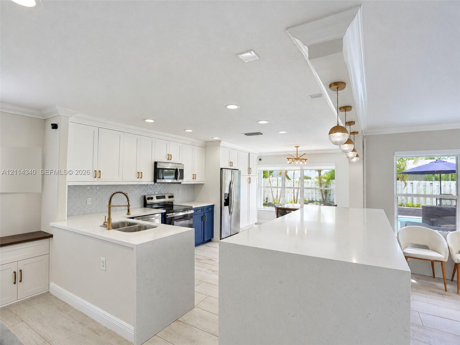 Kitchen featuring an oversized waterfall quartz island, custom white cabinetry with gold-tone hardware, a geometric tile backsplash, stainless steel appliances, and recessed lighting