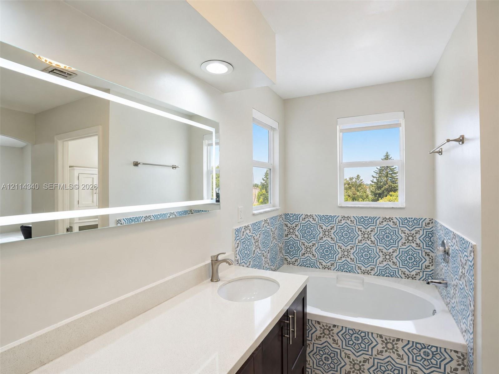 Built-in vanity with white solid-surface countertop and undermount sink, integrated backlit mirror, recessed lighting, and jetted soaking tub featuring patterned ceramic tile surround