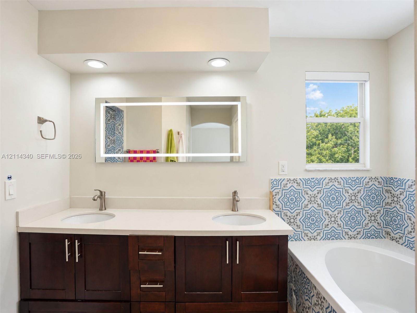 Double vanity featuring a light-toned countertop, dual integrated sinks, dark wood-finish cabinetry, a backlit mirror, and decorative patterned tile surrounding the soaking tub