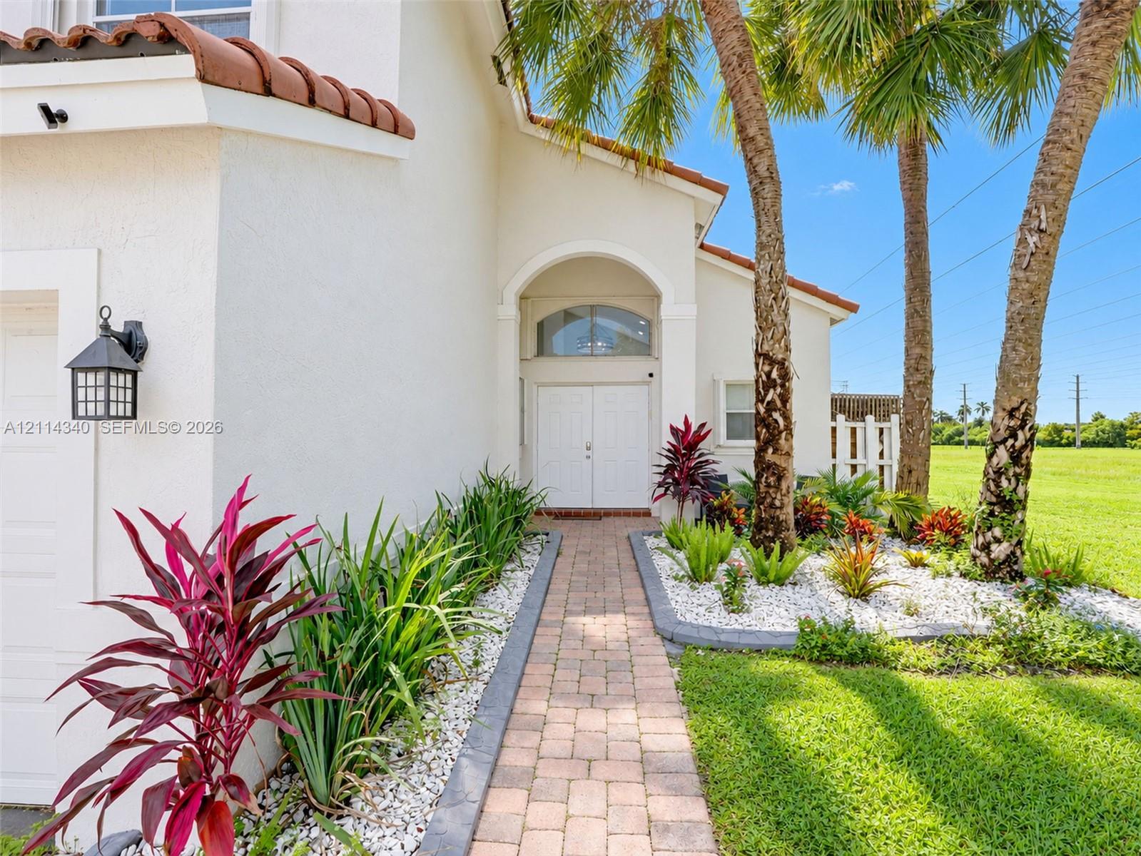 Stucco exterior featuring a tiled roof, arched entryway with double doors, paver walkway, and professional landscaping with mature palm trees