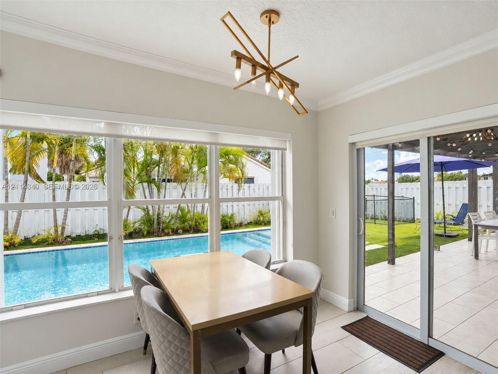 Dining area featuring crown molding, an angular gold-tone multi-light chandelier, large windows, and a glass sliding door providing outdoor access to a tiled patio and swimming pool
