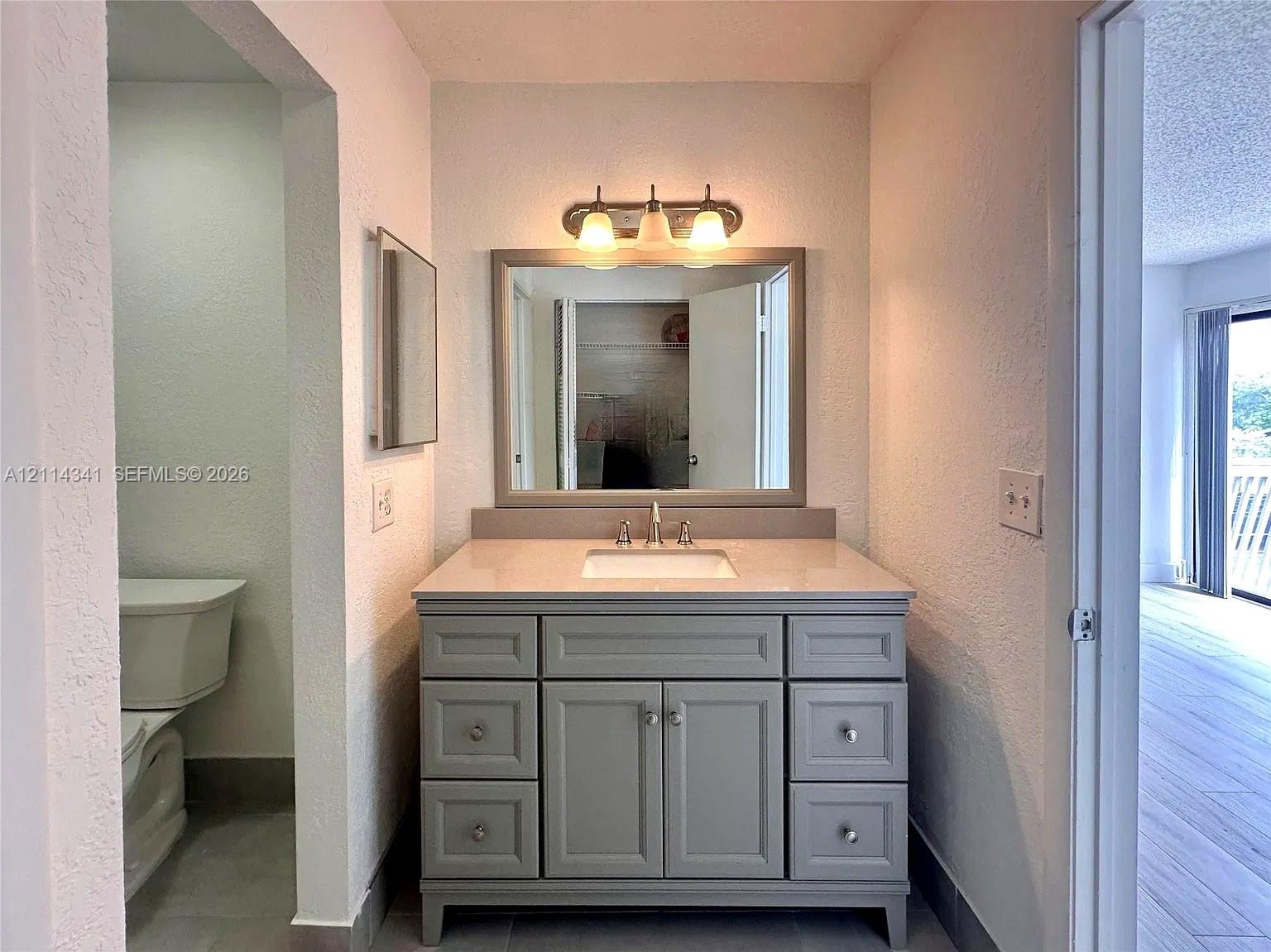 Bathroom vanity featuring a grey cabinet base, light-toned countertop, undermount sink, brushed-finish faucet, and a rectangular mirror with a coordinated frame, illuminated by a three-light fixture Bathroom vanity featuring a grey cabinet base, light-toned countertop, undermount sink, brushed-finish faucet, and a rectangular mirror with a coordinated frame, illuminated by a three-light fixture