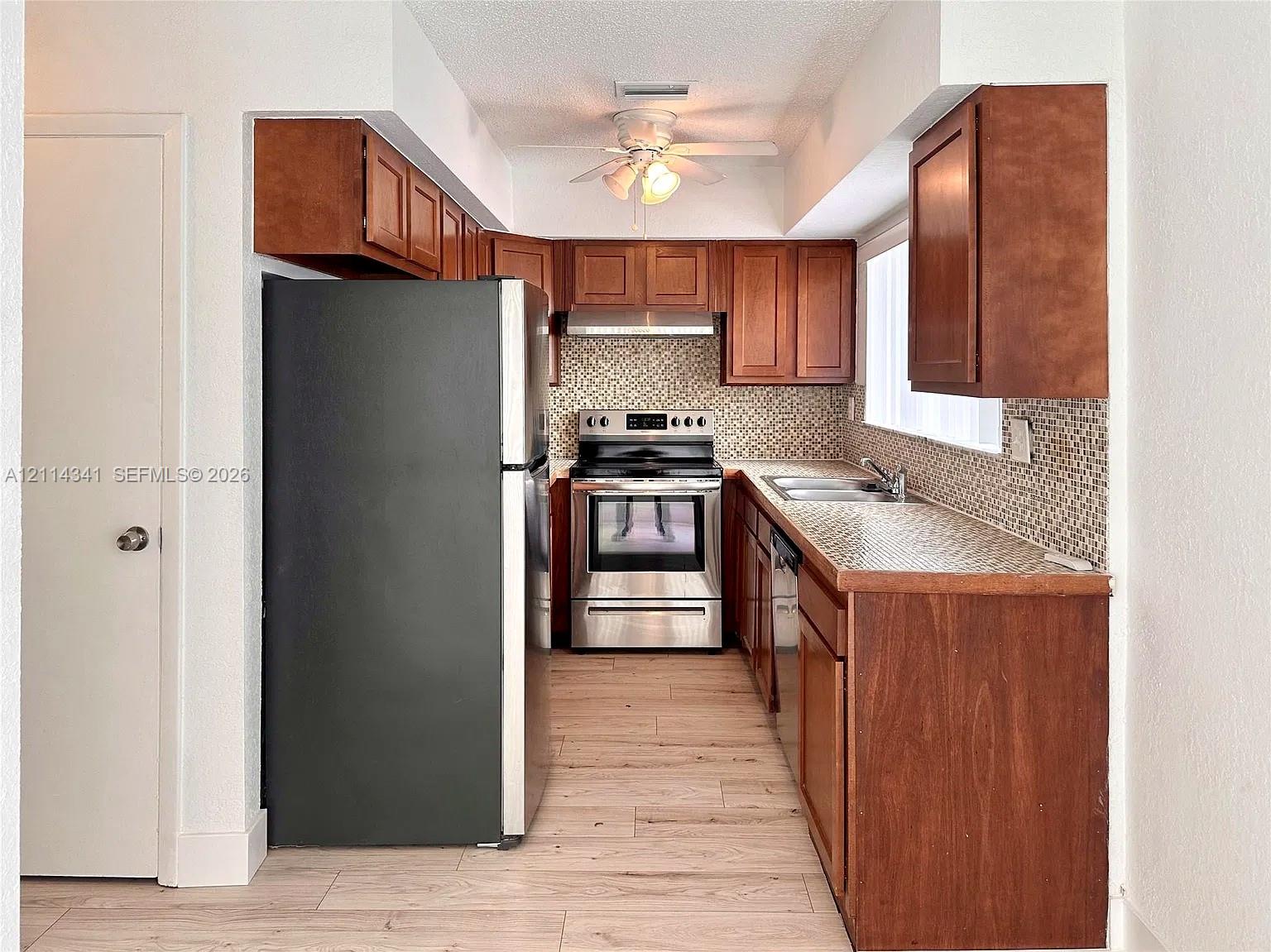 Kitchen featuring wood cabinetry, stainless steel appliances, mosaic tile backsplash, wood-finish flooring, and a ceiling-mounted light fixture Kitchen featuring wood cabinetry, stainless steel appliances, mosaic tile backsplash, wood-finish flooring, and a ceiling-mounted light fixture